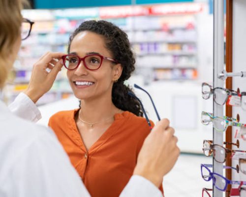 African young woman in optic store choosing new cool-down glasses with optician. Mixed race girl trying new eyeglasses with the help of the pharmacist. Happy multiethnic woman trying new rest spectacle frame in optic store: eyesight and eye care concept.
